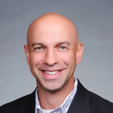 A headshot of Paul Stellato, who is wearing a black suit jacket and plaid shirt.