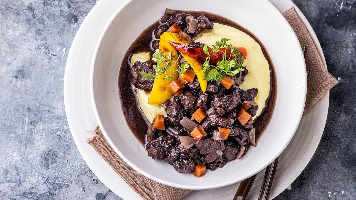 A plant-based boeuf bourguignon, with brown broth and vegetables, is in a white bowl, resting on a white saucer with a beige cloth napkin and wood-handled silverware between the plate and bowl.