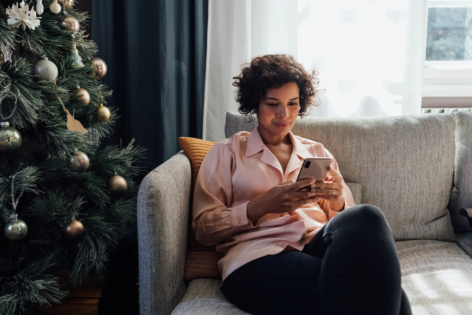 A person sits on a couch holding a mobile phone next to a Christmas tree.