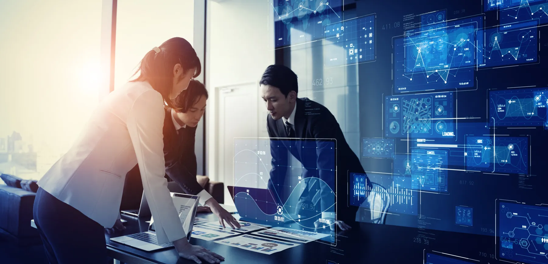 Three people are standing around a table looking at data with digital data on wall.