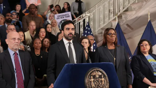 A person wearing a suit speaks into a microphone at a podium surrounded by other people.