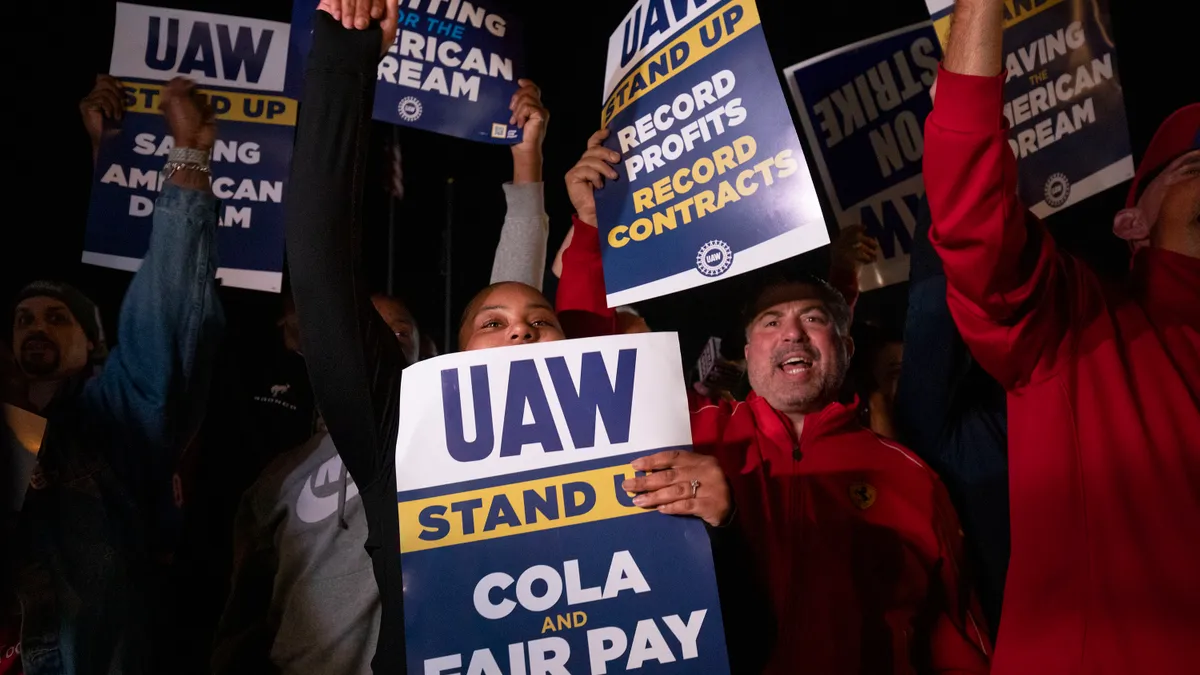 A crowd of striking auto workers at night hold up signs such as Fighting for the American Dream and Stand Up, Record Profits, Record Contracts.