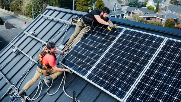 Two people on the roof of a house holding a solar panel. One uses a drill to attach the panel to the roof. Behind the roof are trees and other homes.