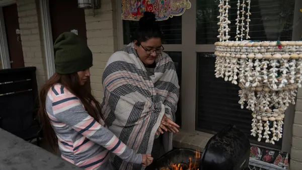 Two people stand next to flames in a barbecue with their hands stretched out over the fire. One person is wrapped in a blanket.