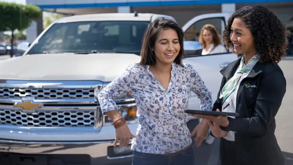 A CarBravo salesperson smiles as she speaks with a potential customer at a GM dealer with a Chevrolet pickup truck in the background.