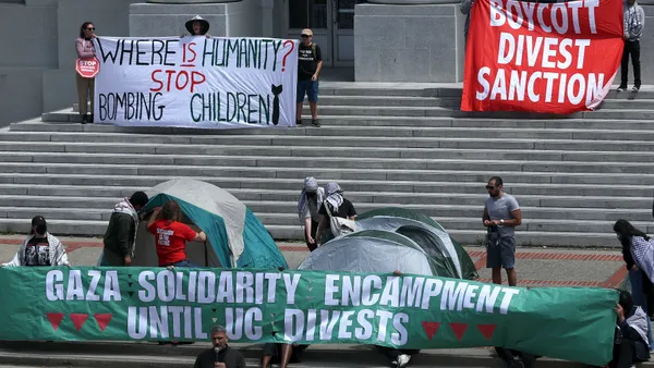 Protestors hold signs that say "Gaza solidarity encampment until UC divests" and "Where is the humanity? Stop bombing children"