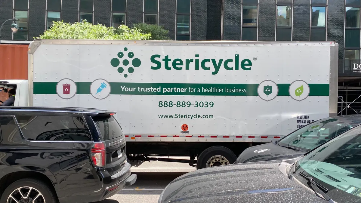 A truck with Stericycle branding sits among cars on the road with a tall office building behind it.