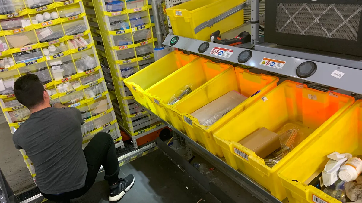 A picker at an Amazon fulfillment center pulls items from shelves atop an Amazon Robotics "drive" base that moves around the cage at the center to be loaded and unloaded.