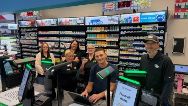 several people stand behind a self checkout machine in a store with a wall of tobacco products behind them. A screen on one of the machines says "Mashgin. Place all items."