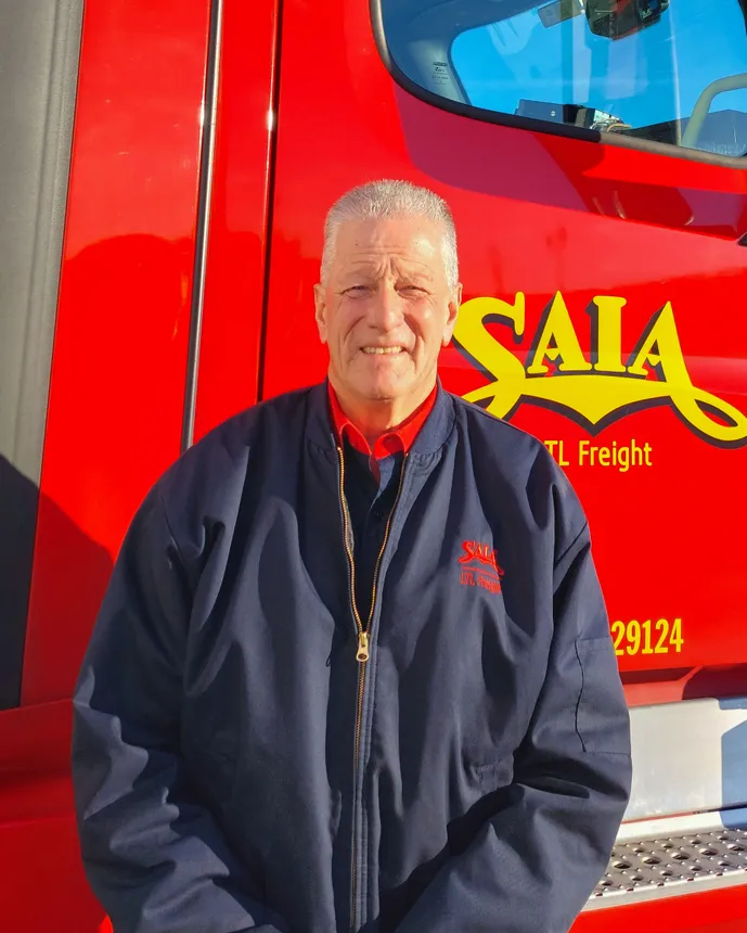 A person smiles in front of his red Saia truck in a navy blue Saia bomber jacket.