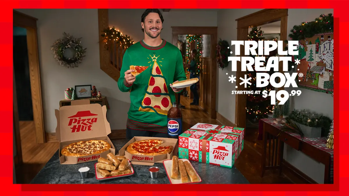 Josh Allen wears a pizza sweater at a holiday party behind a table filled with pizza and Pepsi while holding a slice of pizza.