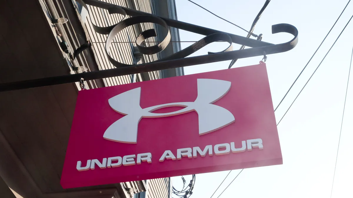 A red sign with a white logo and the words "under armour" in white capital letters hangs from an iron post.