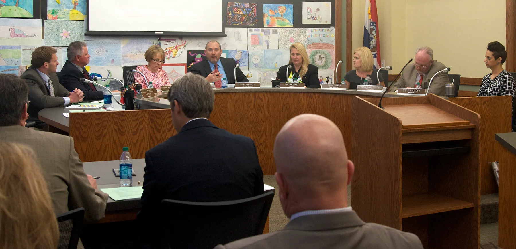 School board members sit during a meeting behind a podium with an audience looking toward them