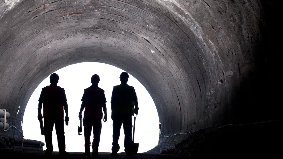 Three construction workers stand in a tunnel, silhouetted in shadow.