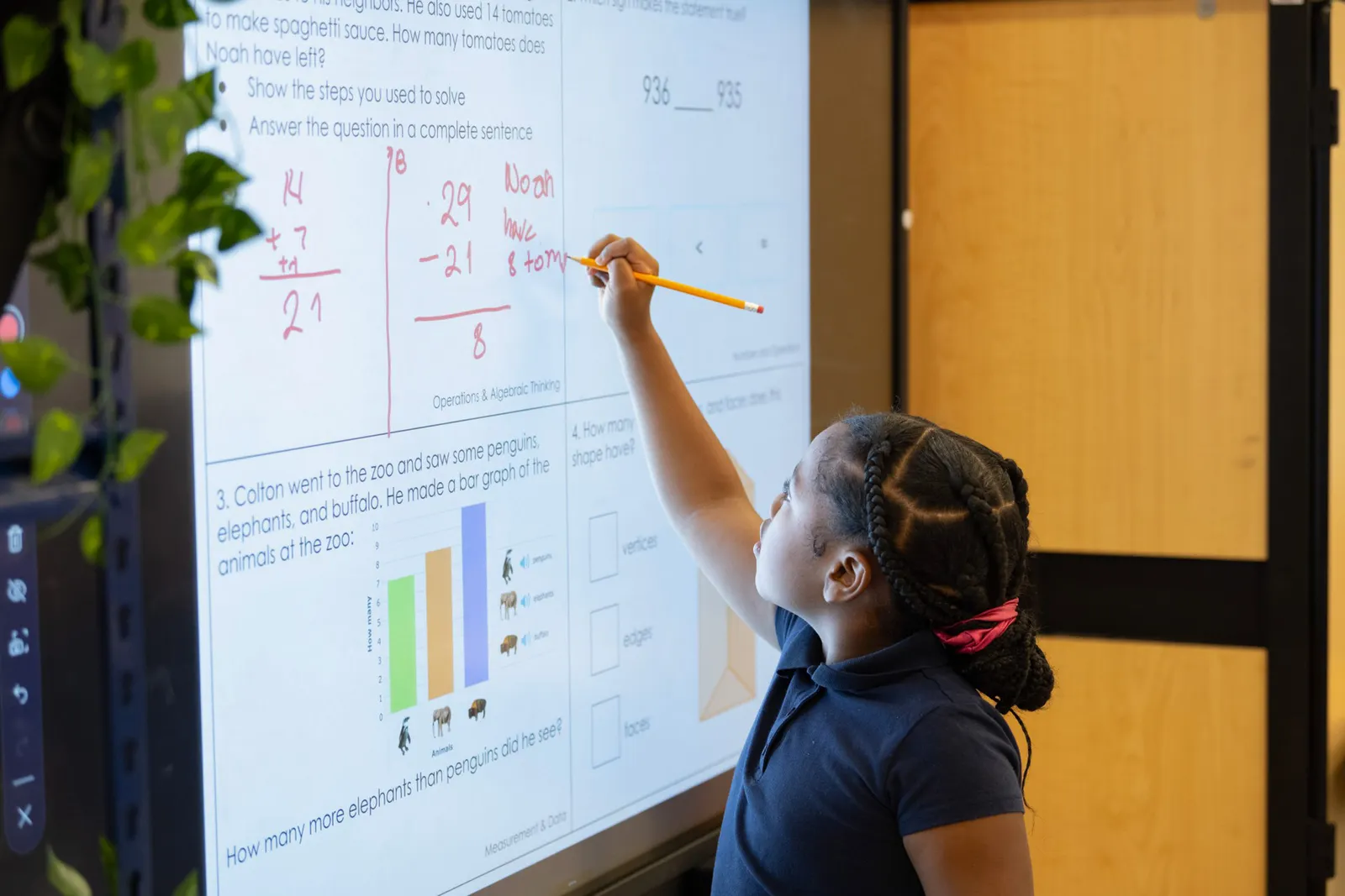 A young student is facing a smart board in a classroom and is using a writing tool to work on math problems on the board.