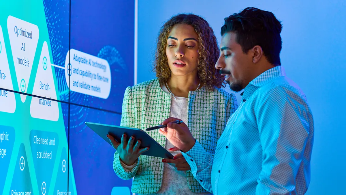 Two people in a blue-lit room discuss data on a tablet. A large digital display with AI-related keywords is in the background. The setting is futuristic and focused.