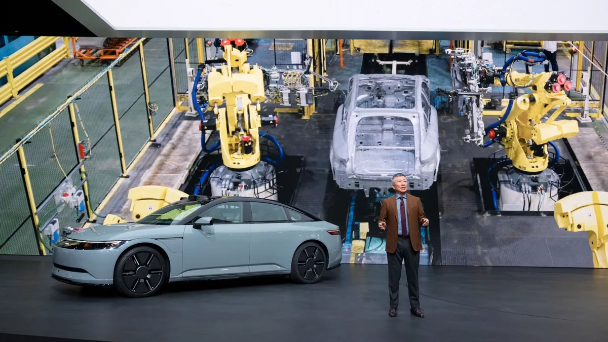 Sony Honda Mobility's Yasuhide Mizuno speaks on a stage next to a prototype Afeela vehicle, while a projector shows an image of the vehicle's manufacturing process.