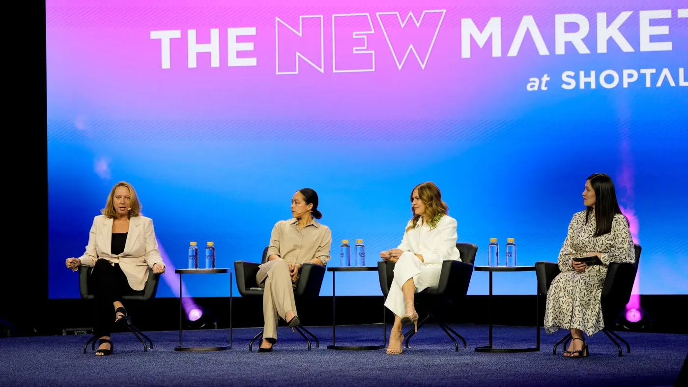 Four people sit in chairs on a stage with the words "The New Market at Shoptalk" behind them.