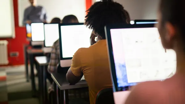 Students work on computers while a teacher instructs in front of them in a classroom.