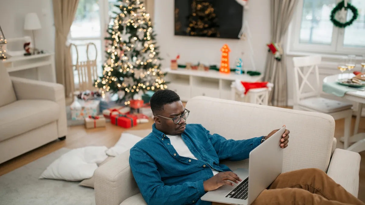 Person sits on an oversized chair with a computer and a Christmas tree, presents and Santa hat in the background.