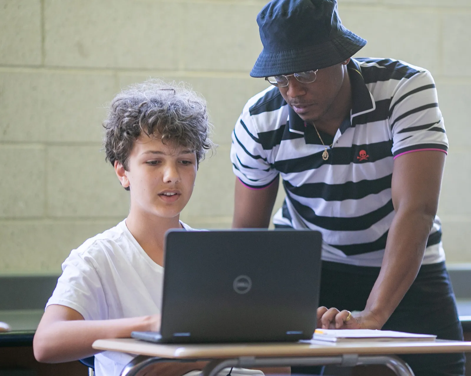 A student sits at a desk looking at an opened laptop. An adult to to the students' left-hand side and is looking at the laptop.