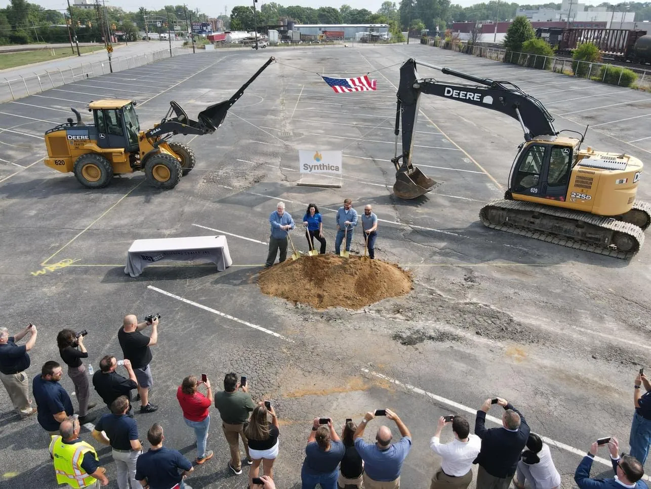 Overhead view of four people shoveling dirt in a ceremony on a dirt industrial site with photographers looking on.