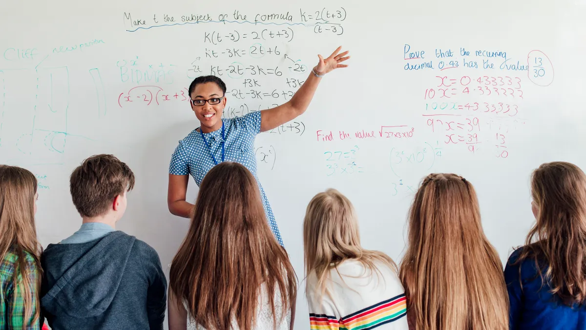 A teacher instructs a math class in front of a white board with written equations.