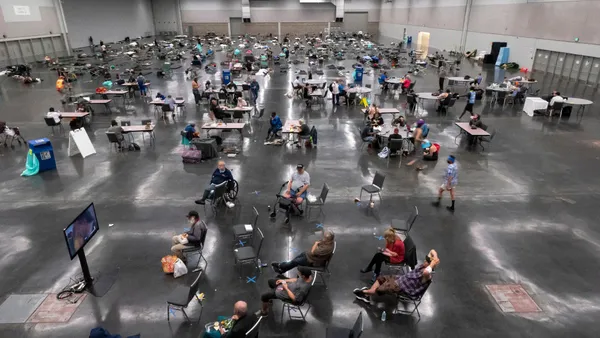 Aerial shot of people inside a large indoor space. People sit on folding chairs and lay on mats on the floor.