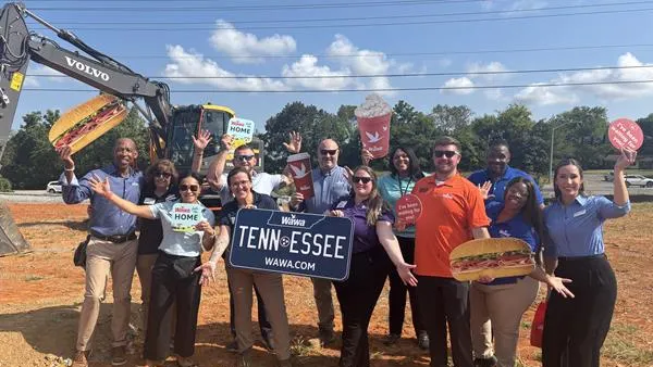 A photo of a number of people standing on a construction site. There is a Volvo backhoe in the background. The people are holding up signs showing food items and one that looks like a license plate that reads "Wawa Tennessee wawa.com."