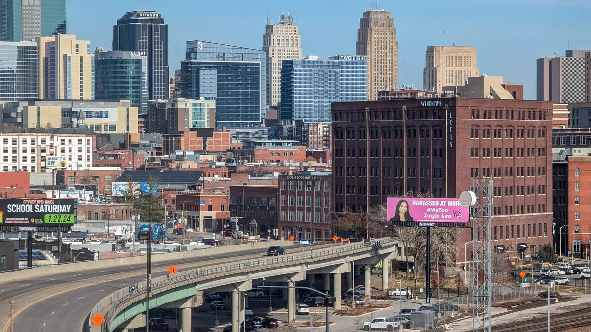 A highway and railroad tracks cross in front of tall brick buildings.