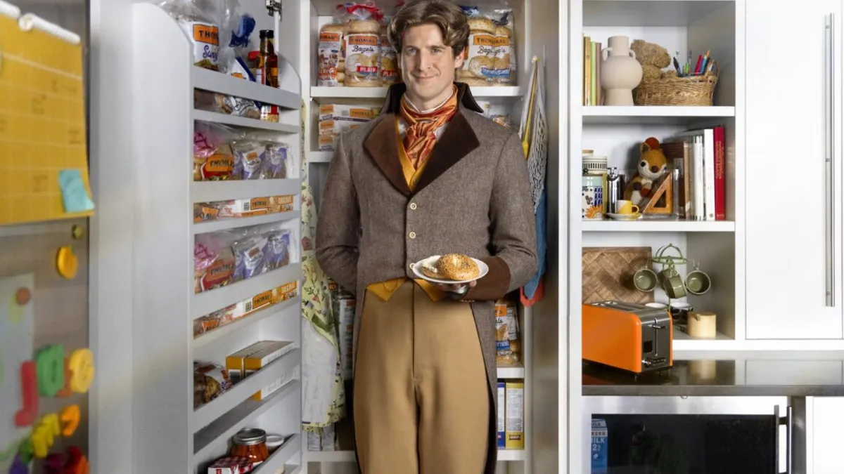 An image of Thomas' Breakfast's new brand character, Thom, standing tall in a kitchen pantry and holding out a plate of bagels.