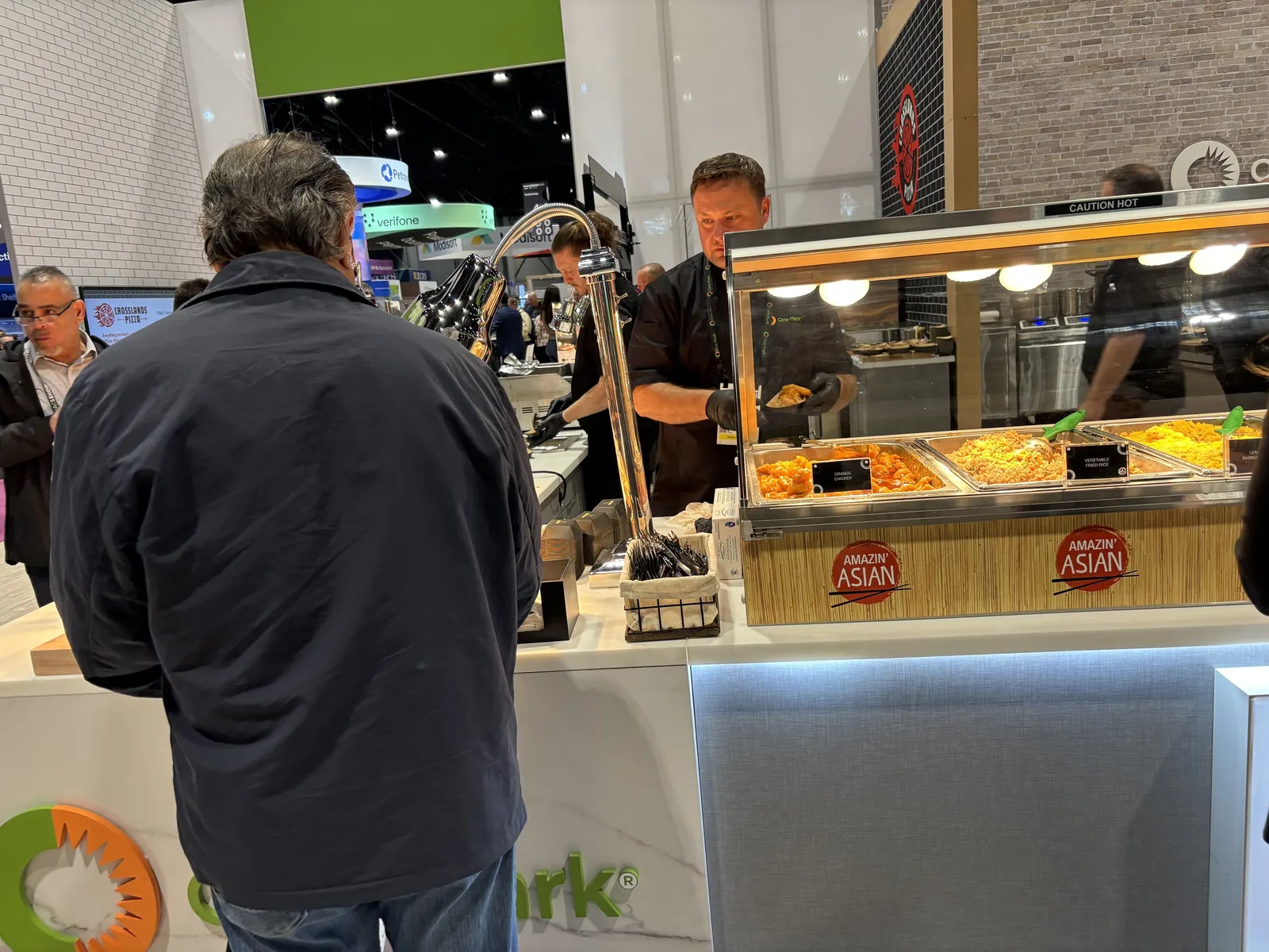 A photo of people getting food at a book at a trade show. Signs on the front of a heated case full of food say "Amazin' Asian" and "orange chicken" and "Vegetable fried rice."