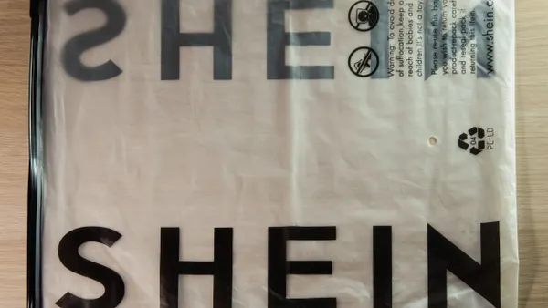 A clear plastic bag with large black text reading "SHEIN" printed on it. The bag is laying flat on a light wooden surface, with a black zipper strip on the left edge. Small warning text and symbols are printed in the upper right corner.