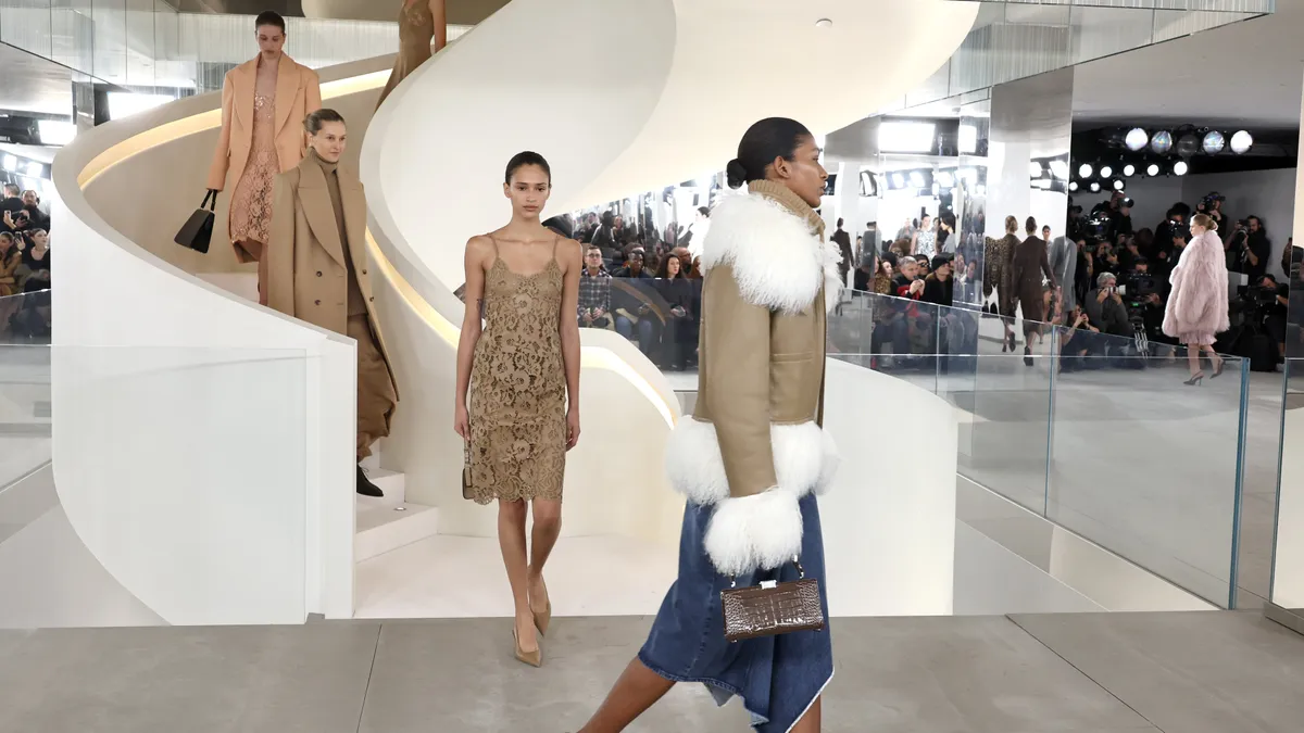 Models walk down a curved white staircase wearing camel-colored outfits and one model crosses in front of the camera in a camel-colored jacket and blue denim skirt.