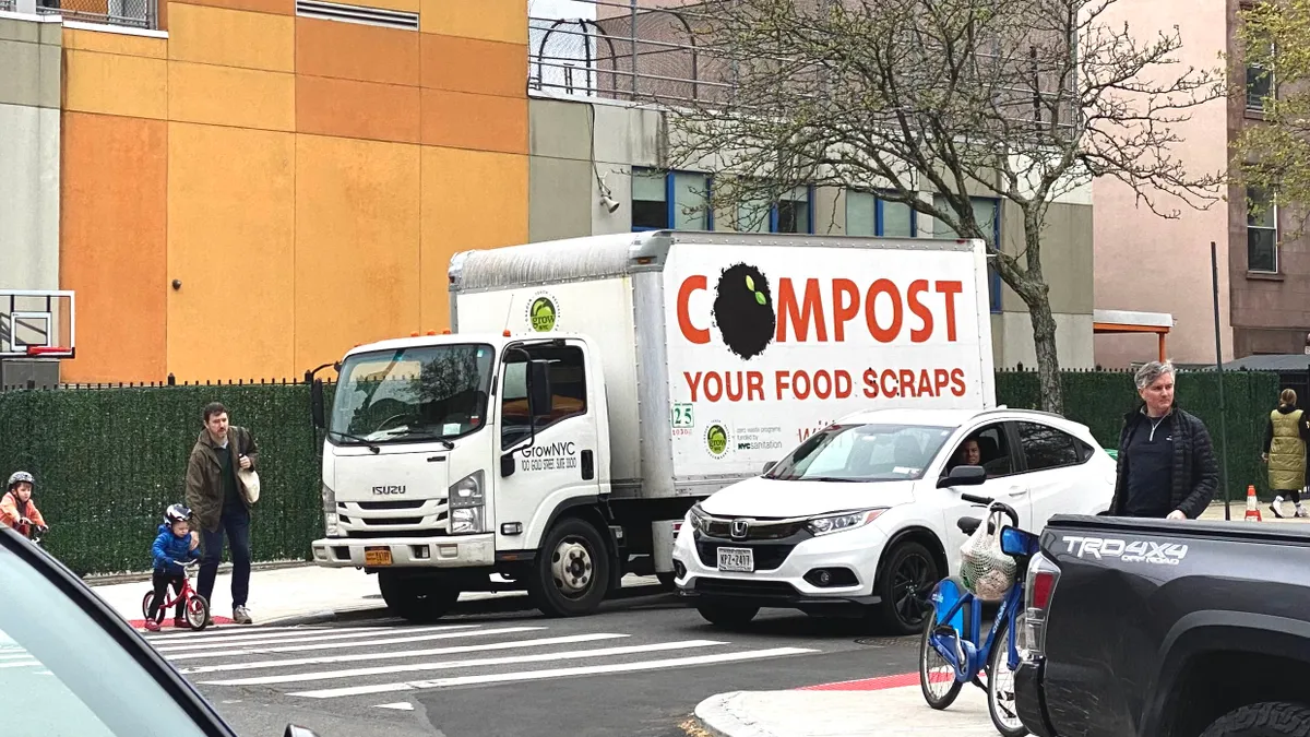 A truck with large letters that read "Compost your food scraps" and has GrowNYC logos is parked near a building and crosswalk.