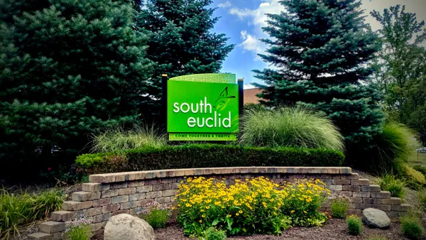A green welcome sign outside surrounded by pine trees and hedges.