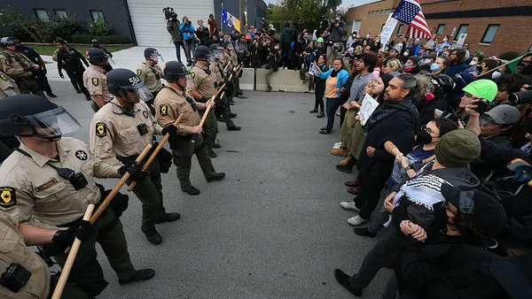 two groups of people are standing on a street facing each other. One side has posters and the other side is dressed in law enforcement uniforms.