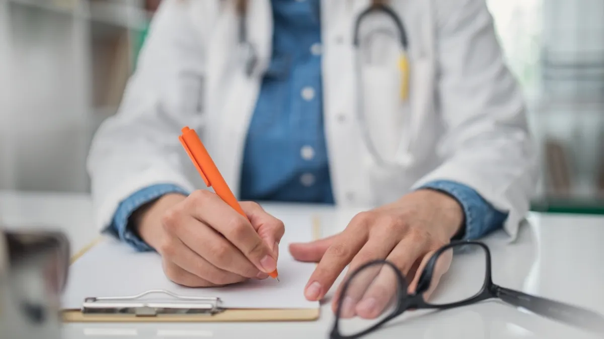 Doctor sitting down at desk, writing on clipboard with glasses to the side.
