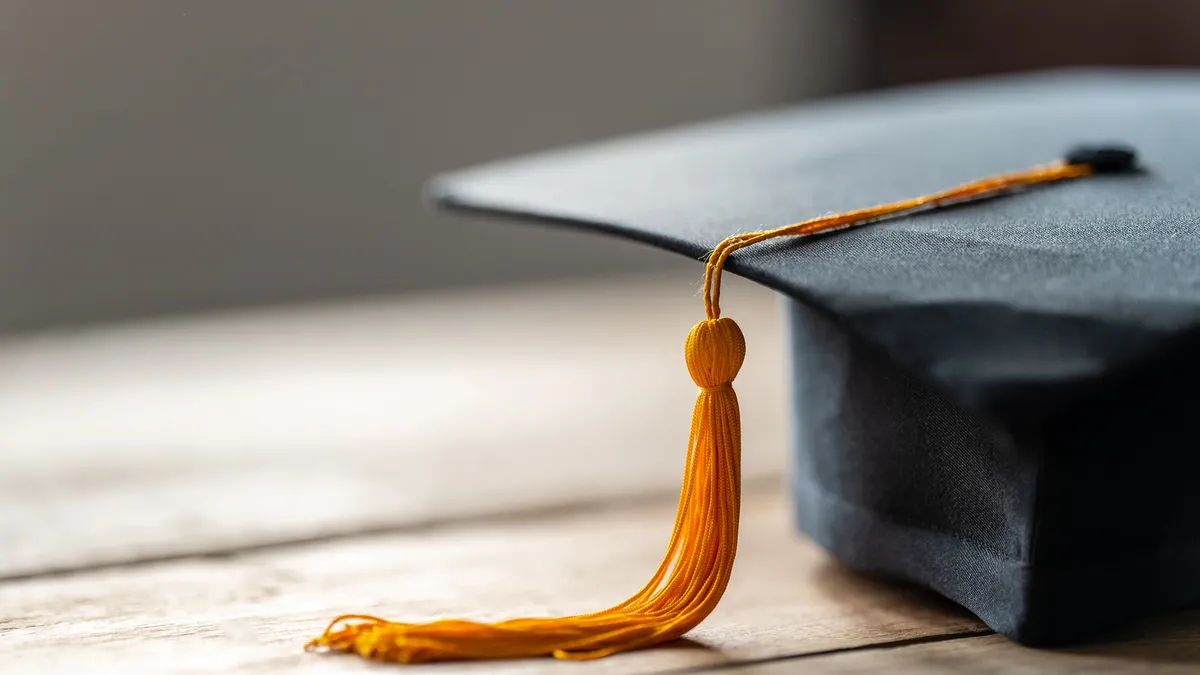 A close-up photo of a  black graduation cap with a yellow tassel hanging at the left side sits on a wooden table.