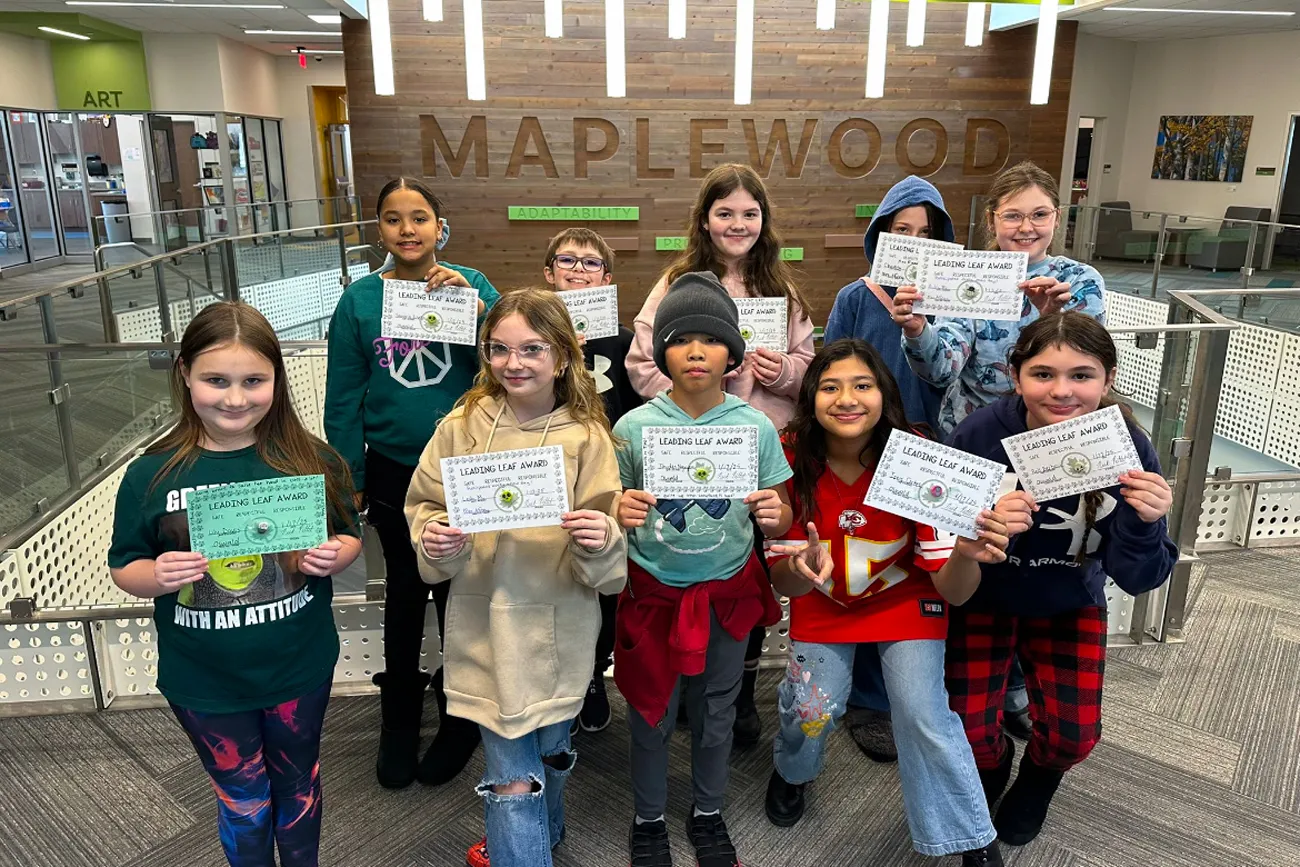 A group of students are gathered standing in a school hallway near stairs. They are holding up certificates and many are smiling.