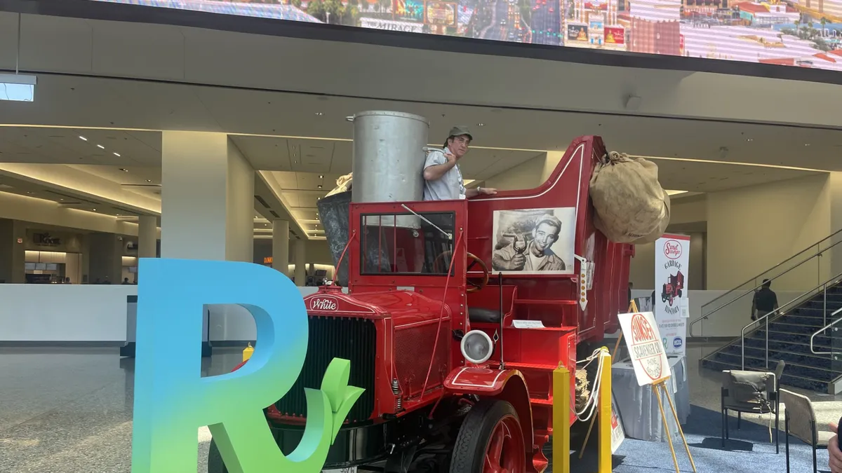 Person holding an old garbage can standing on top of a red antique garbage truck