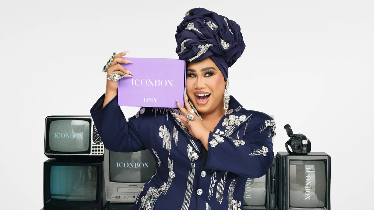 Patrick Starrr holding an Ipsy Icon Box standing in front on televisions.