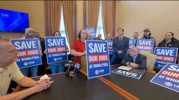 A woman standing behind a podium with people holding up blue, white and red "Save our jobs" signs standing behind her.