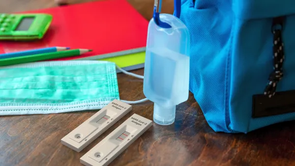 A backpack sits on a wooden desk. Attached to backpack is hand sanitizer. Also on the desk are COVID-19 rapid tests, a mask and notebooks