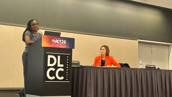 A person stands at a podium while speaking. Another person is sitting to the right at a table.