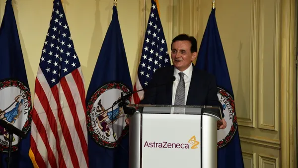 A person in a dark business suit speaks at a podium in front of U.S. and Virginia flags.