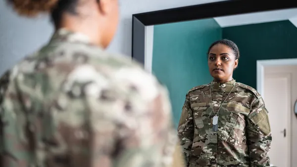 A Black woman, in military uniform, contemplates her reflection in the mirror