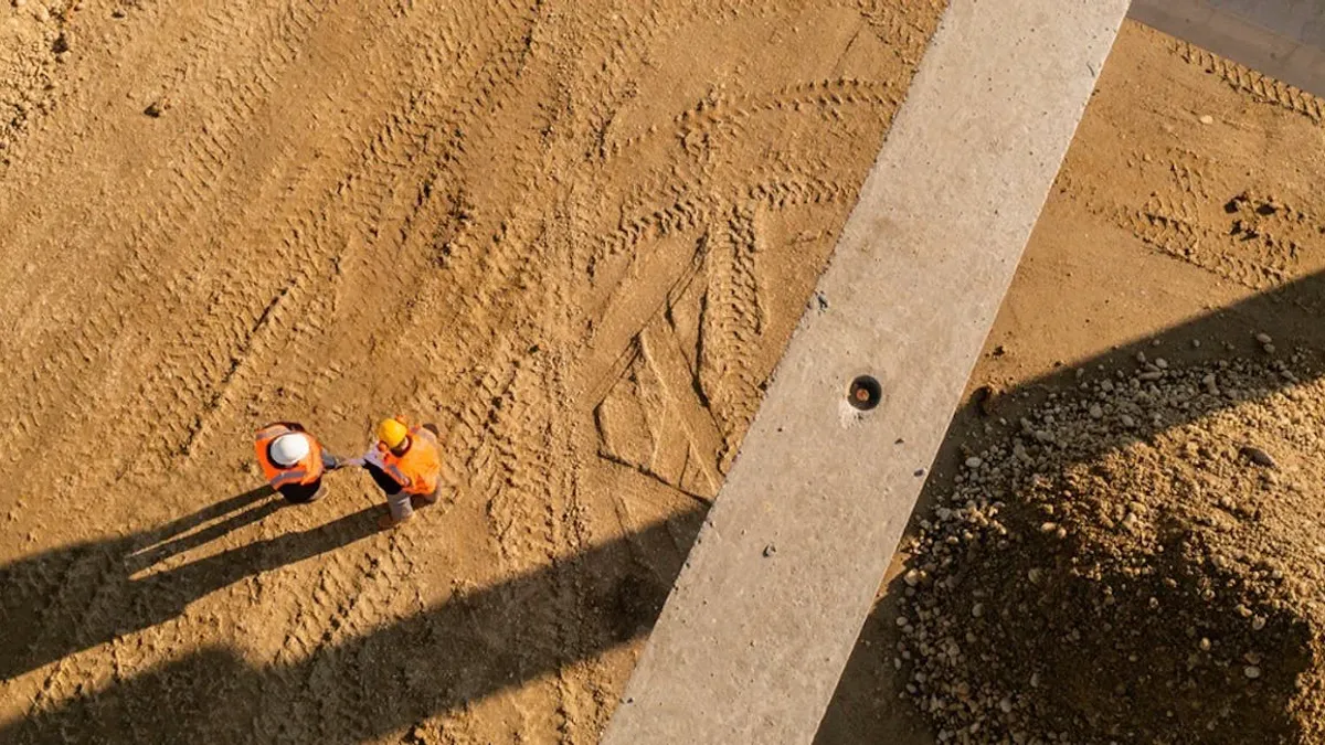 Two workers stand on a field of dirt, while the shot is staged from directly overhead, elevated in the air.