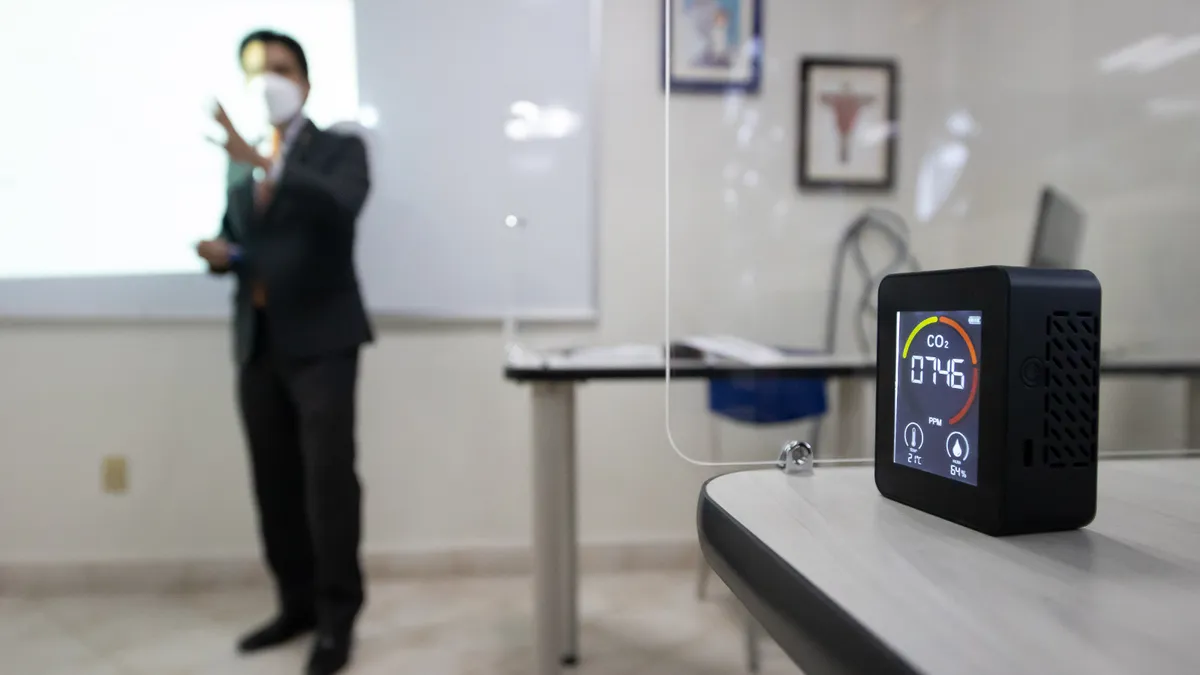 A teacher instructs in a room, in the foreground a carbon dioxide meter is used to measure air quality and ventilation.