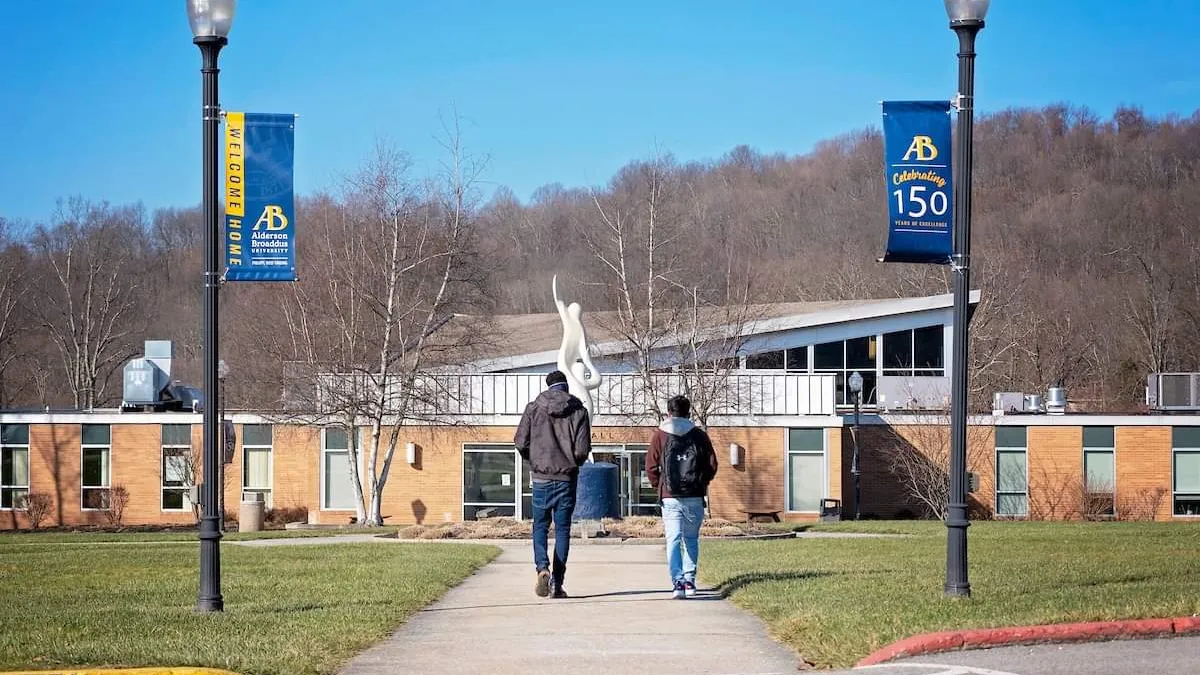 Two people walk on the Alderson Broaddus campus, with light posts next to them with signs advertising for the university.